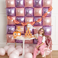 Smiling girl in pink overalls poses beside trick-or-treat table topped with stacked pumpkin boxes and candy, against shimmering balloon wall and hanging fans.