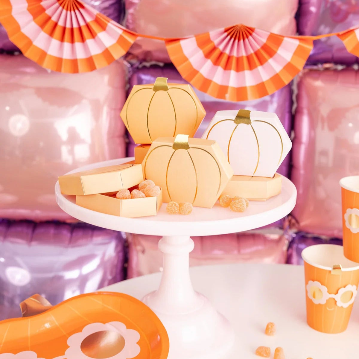 Bright party tablescape with pumpkin favor boxes, orange sunglasses plate, matching cups and scalloped napkins in front of pink and purple balloon wall backdrop.