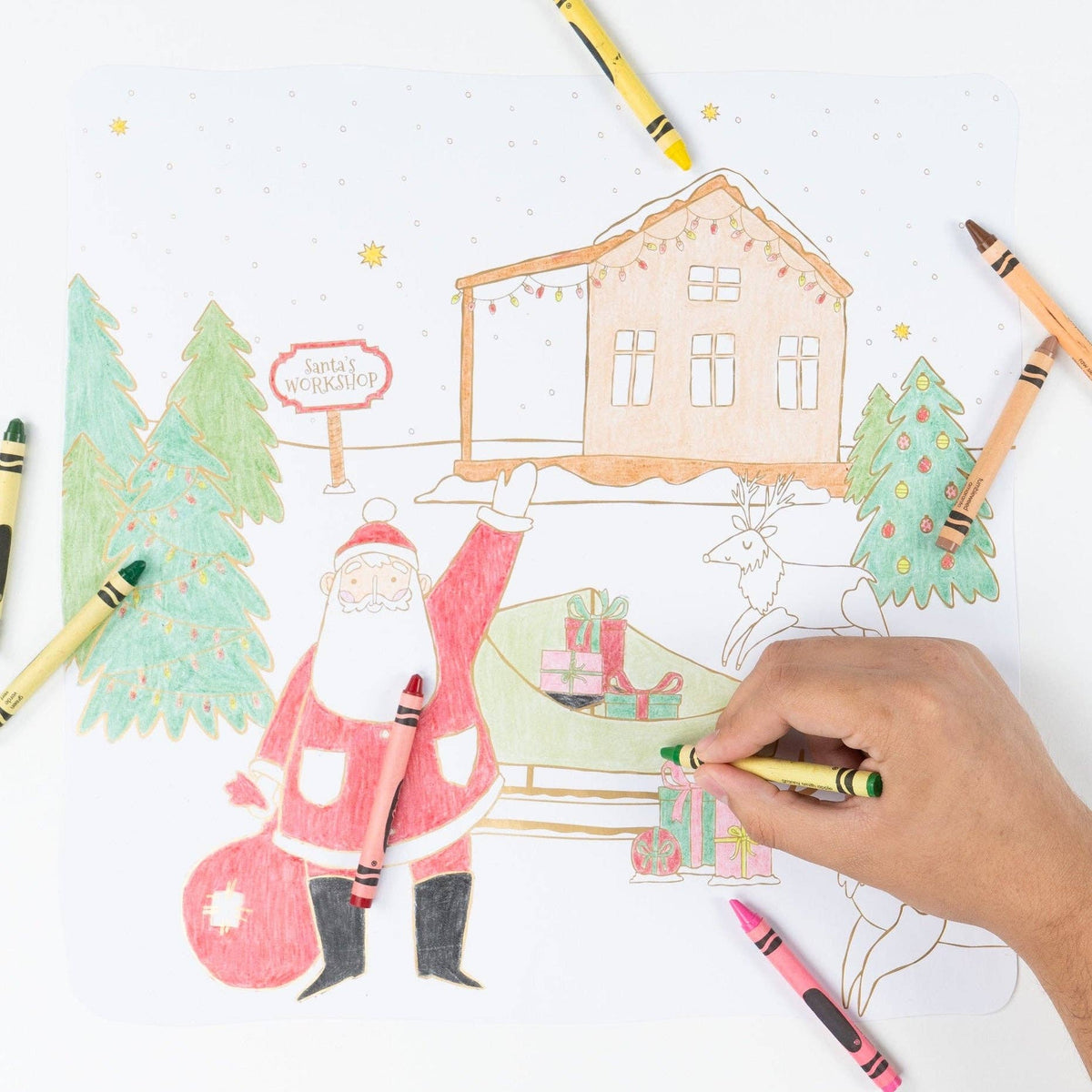 Wide shot showing hands coloring the winter village scene with crayons and markers.