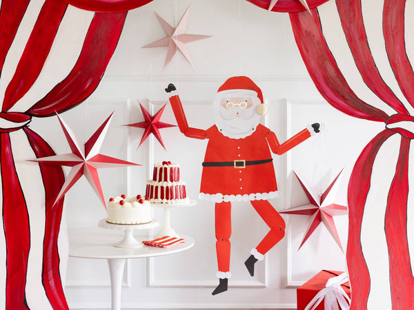 The jointed Santa hanging on a red-and-white striped backdrop beside 3D paper stars above a dessert table.