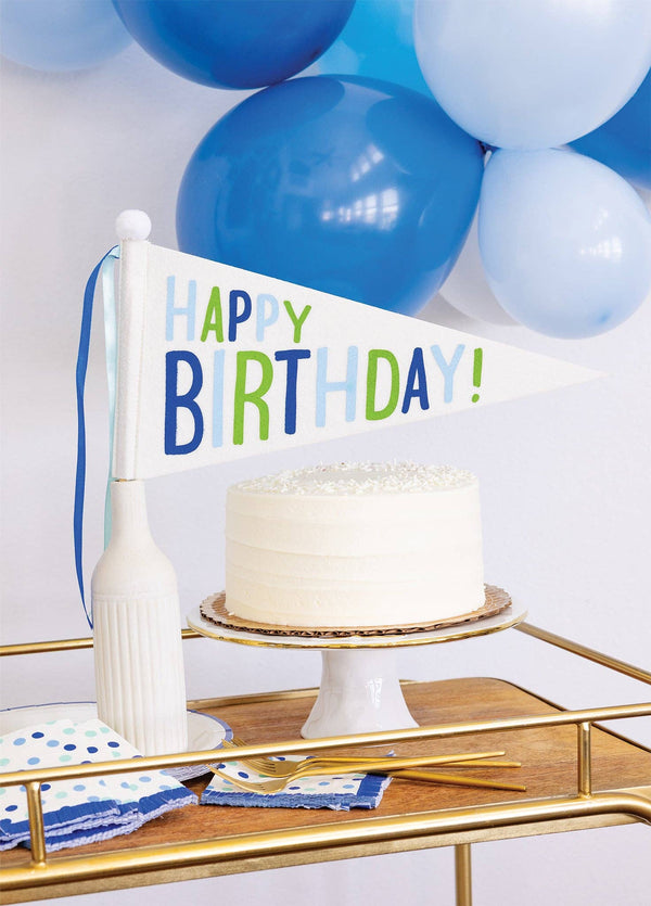 Happy Birthday blue and green felt pennant flag displayed next to white birthday cake on gold bar cart with blue balloons