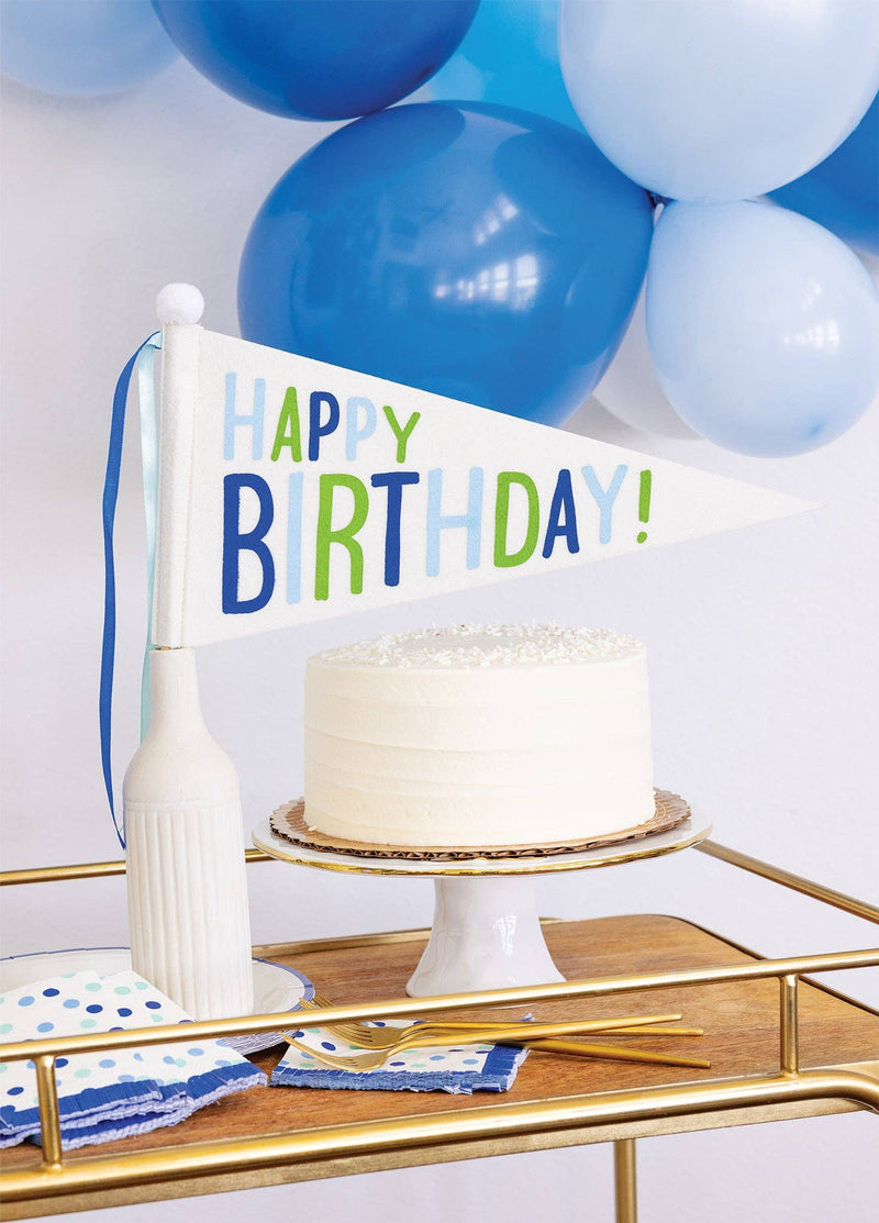 Happy Birthday blue and green felt pennant flag displayed next to white birthday cake on gold bar cart with blue balloons
