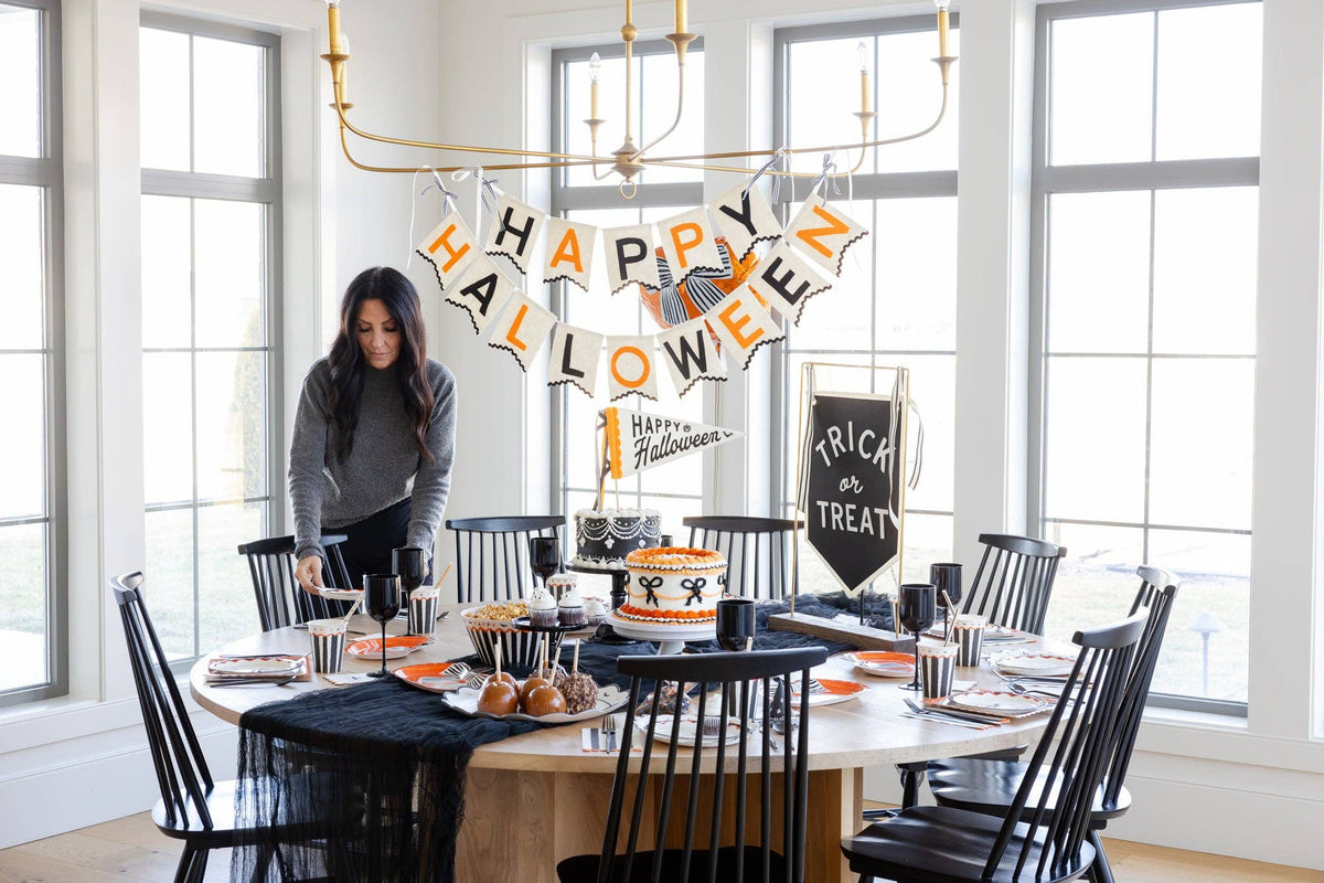 Woman setting a Halloween table beneath a Happy Halloween felt banner, with black and white striped cups, orange plates, and two decorated cakes.