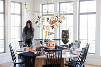 Woman setting a Halloween table beneath a Happy Halloween felt banner, with black and white striped cups, orange plates, and two decorated cakes.