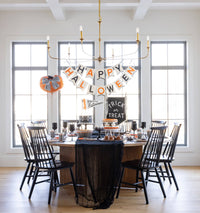 Happy Halloween felt banner in black and orange letters on cream felt, hung beneath a brass chandelier over a round Halloween tablescape with black chairs and a black gauze runner.