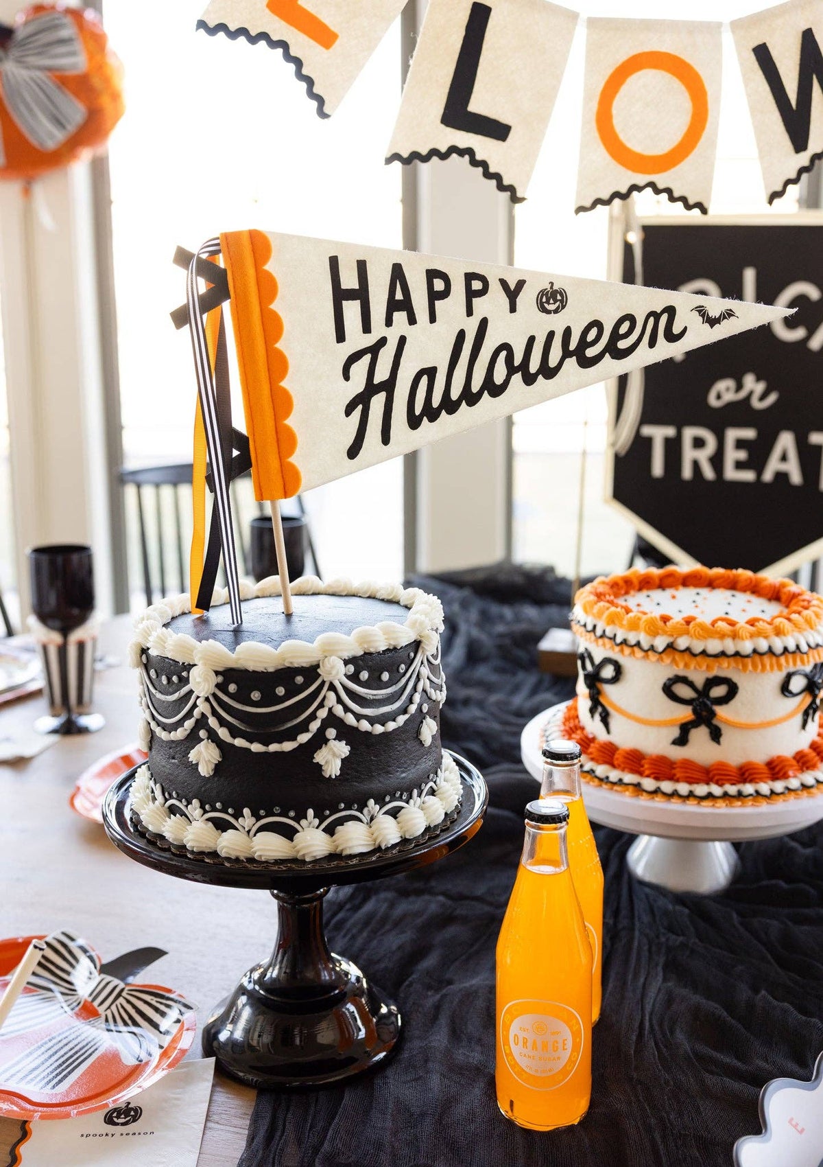 Close-up of the Happy Halloween felt pennant as a cake topper between a black draped cake and an orange bow cake, with orange soda bottles and a Trick or Treat sign.