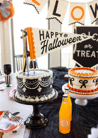 Close-up of the Happy Halloween felt pennant as a cake topper between a black draped cake and an orange bow cake, with orange soda bottles and a Trick or Treat sign.
