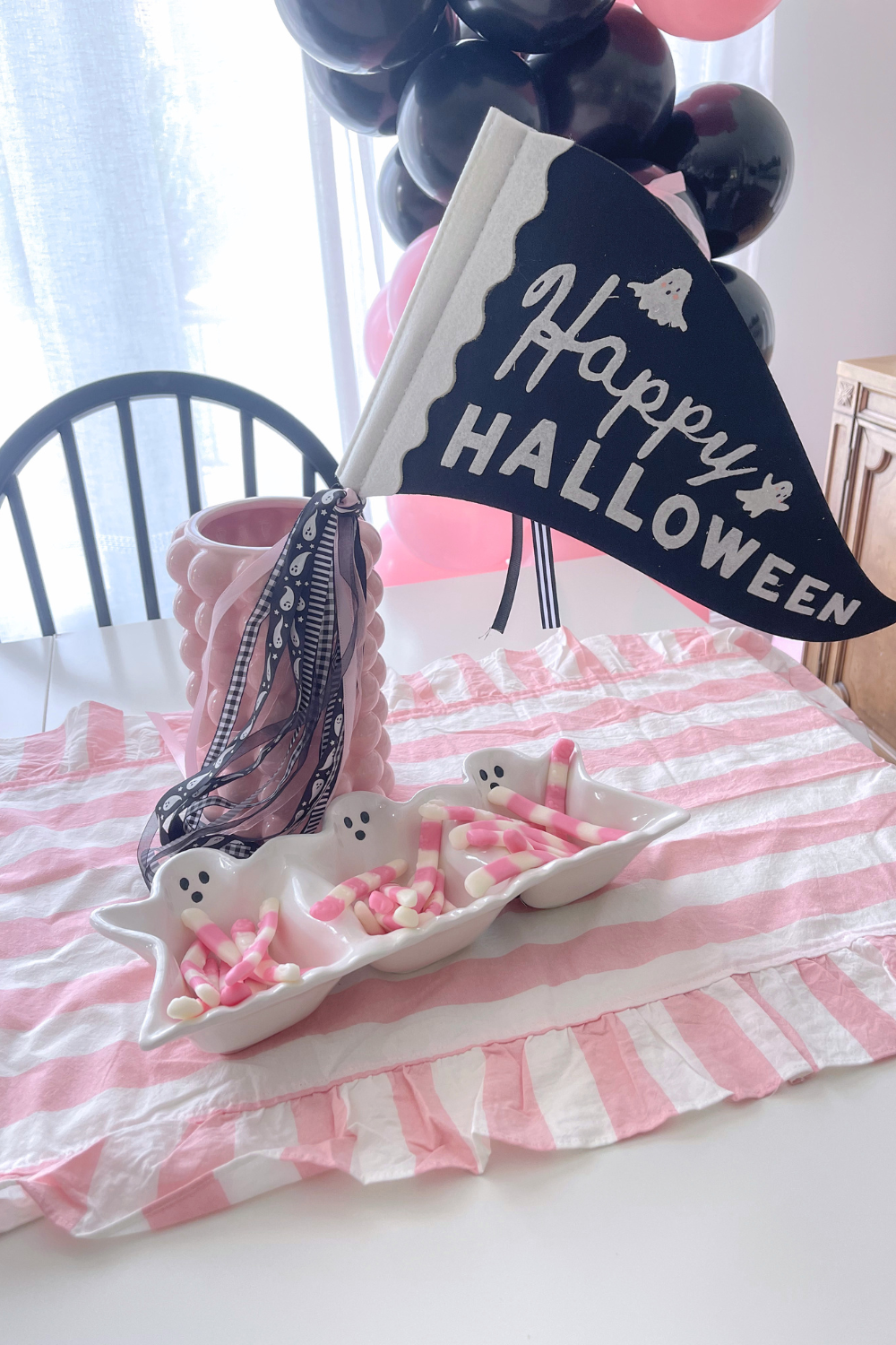 Pink Halloween party table with a navy "Happy Halloween" ghost pennant flag, a white ghost-shaped divided candy dish holding pink and white candy, a pink scalloped vase with ghost-print ribbon, a pink and white striped ruffled runner, and a black and pink balloon garland