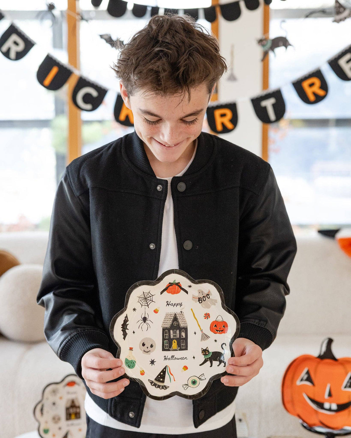 A boy in a black bomber jacket holds the plate at chest height, looking down at it. Full plate face visible: cream background, black scalloped edge, haunted house center, surrounded by pumpkin, ghost, spider, bat, cat, skull, potion bottle, witch hat, eyeball, candy corn, and "Happy Halloween" script. Trick or Treat felt banner and character garland visible in background.