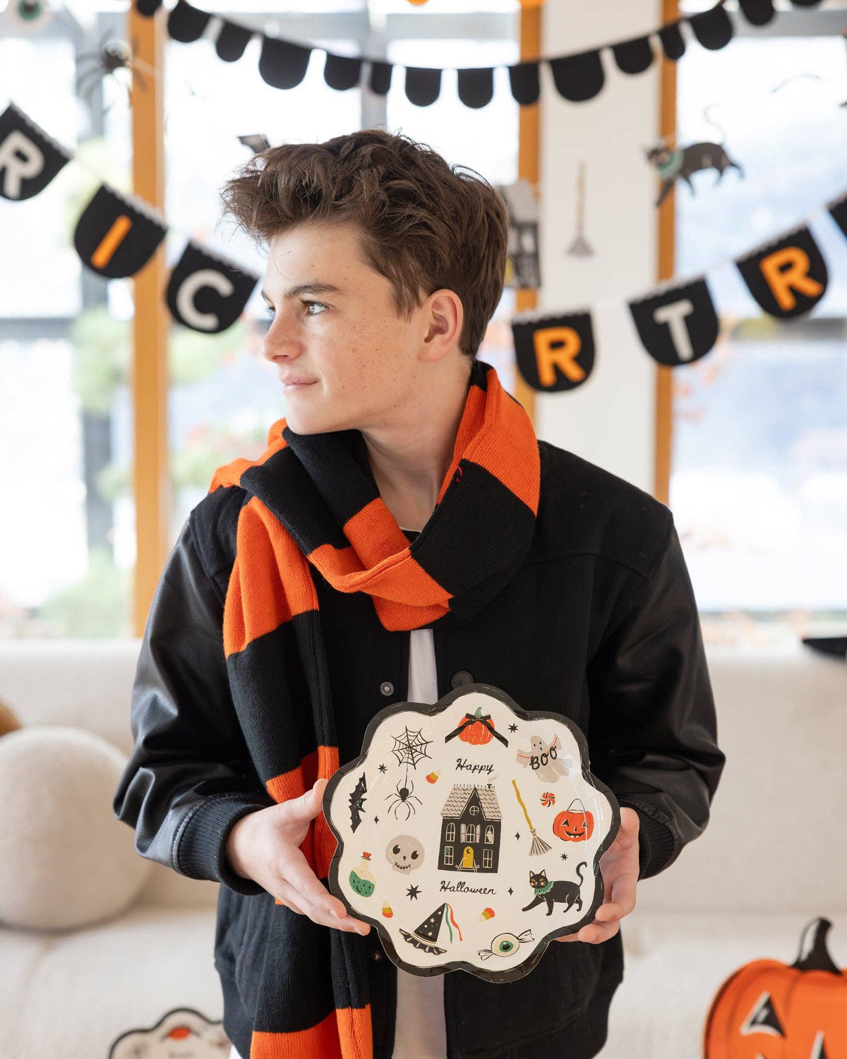 A boy wearing a black-and-orange striped scarf holds the plate at waist height, looking to the side. Full plate illustration visible. Trick or Treat felt banner and character garland in background. Pumpkin-shaped plates visible at lower right edge.