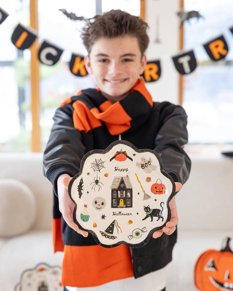 Boy in black jacket and orange scarf holds the plate directly toward the camera, arms extended. Full plate face in sharp focus. Trick or Treat banner and character garland blurred in background. Pumpkin-shaped plates visible at lower right.