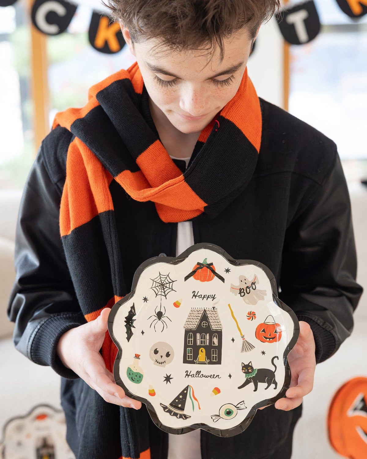 Close-up of the same boy holding the plate at chest level, looking down. Detailed view of the plate's illustrations: haunted house with yellow door and gray scalloped roof, orange pumpkin with black bow, ghost with "BOO" banner, spider on web, potion bottle, skull with stitched smile, black cat with teal collar, witch hat with streaming ribbons, winged eyeball, candy corn, broom.