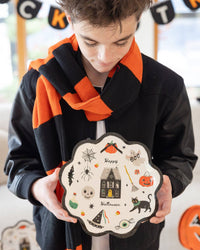 Close-up of the same boy holding the plate at chest level, looking down. Detailed view of the plate's illustrations: haunted house with yellow door and gray scalloped roof, orange pumpkin with black bow, ghost with "BOO" banner, spider on web, potion bottle, skull with stitched smile, black cat with teal collar, witch hat with streaming ribbons, winged eyeball, candy corn, broom.