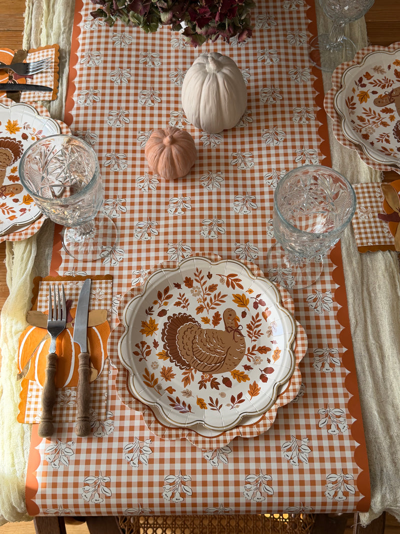 Orange gingham scalloped paper plates layered under fall turkey plates along a gingham floral table runner with pumpkins and crystal goblets