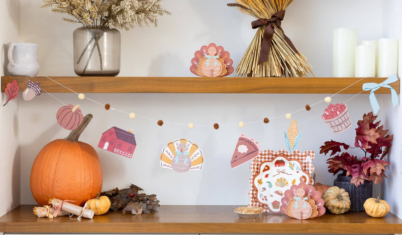 Harvest pom pom garland draped across a styled shelf with pumpkins, candles, and autumn-themed decorations on a white wall.