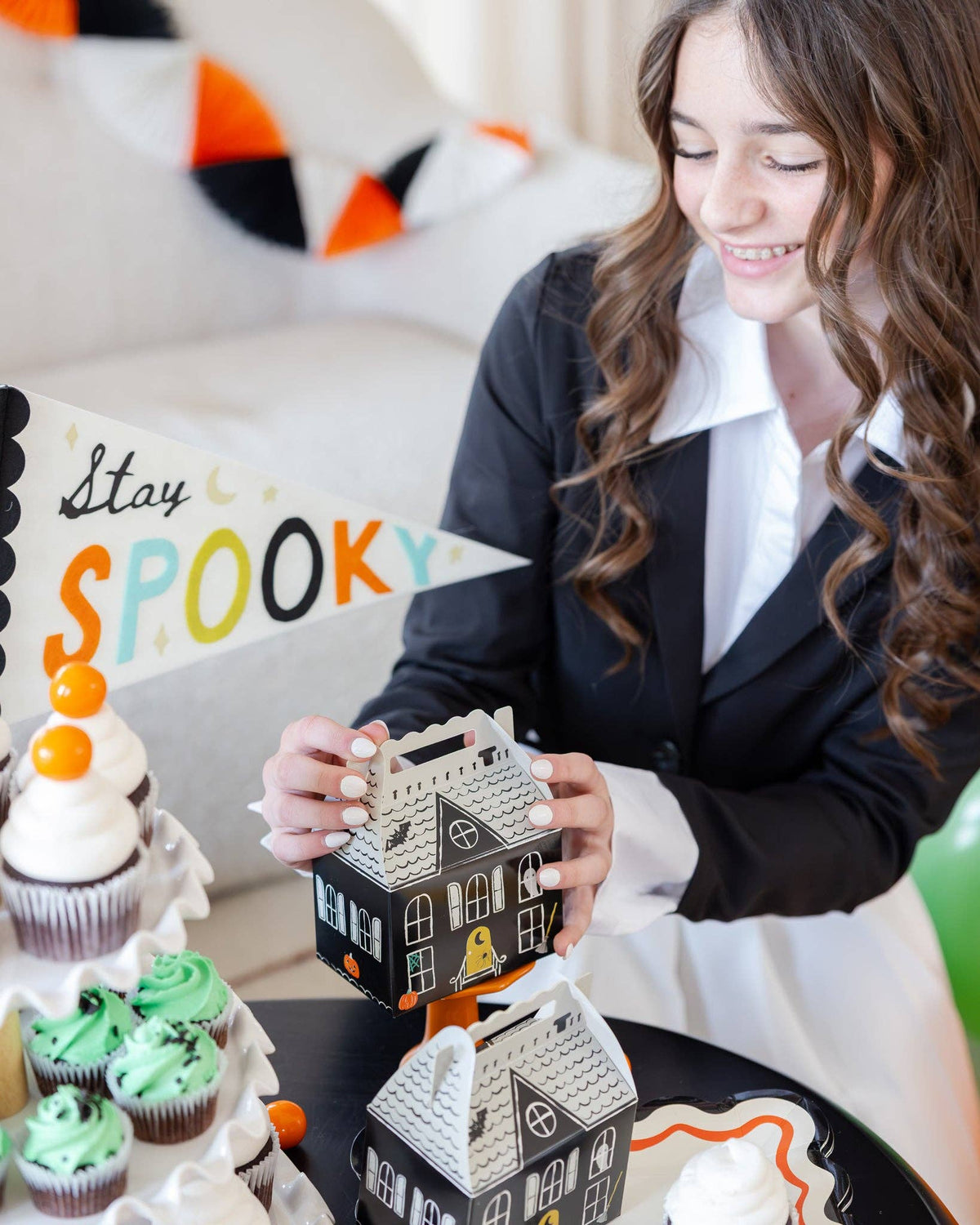 Teen girl looking down and smiling while holding an assembled box; a "Stay Spooky" pennant sits at left, an orange/black/cream felt tassel garland drapes the sofa behind, and cupcakes plus a second box on an orange pedestal anchor the foreground. Lifestyle shot.