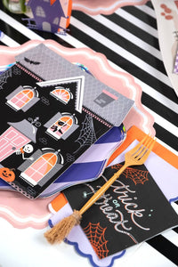 Close-up of a single haunted house Halloween paper plate with a gold fork and a black trick or treat card on a black and white striped surface