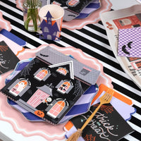 Overhead Halloween party table with haunted house paper plates, gold forks, pink scalloped chargers, and purple accents on a striped runner