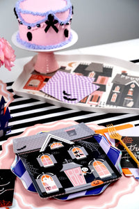 Halloween dessert display with a black bow cake on a stand, haunted house paper plates, and purple gingham details