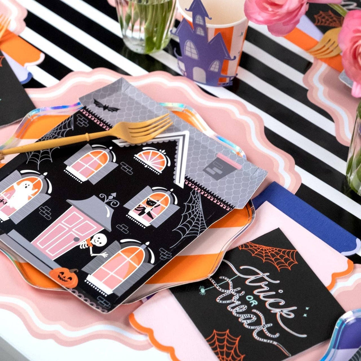 Halloween tablescape with haunted house shaped paper plates, pink scalloped plates, a black trick or treat script card, and pink roses on a black and white striped runner