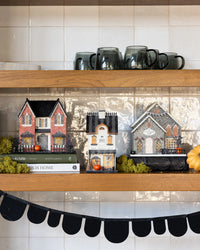 Three glitter Halloween village houses on a kitchen shelf with books, moss and a white pumpkin below a black scalloped banner