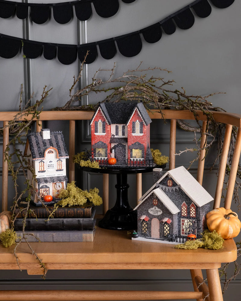 Red Victorian haunted house raised on a black cake stand with two glitter Halloween village houses, mossy branches and pumpkins
