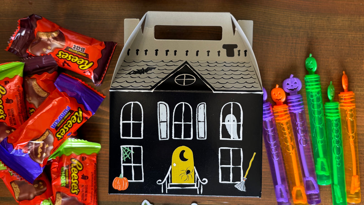 Single white haunted house paper treat box with a carry handle beside a row of rainbow crayons on a wood table
