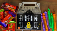 Single white haunted house paper treat box with a carry handle beside a row of rainbow crayons on a wood table