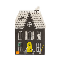 A single haunted house–shaped napkin on a white background: tall die-cut black house with a cream wave-shingled roof and chimney, arched lit windows, a ghost in an upper window, a bat, green spiderweb, broom, orange pumpkin, and a yellow front door with a crescent moon and spider. Clean overhead product shot.