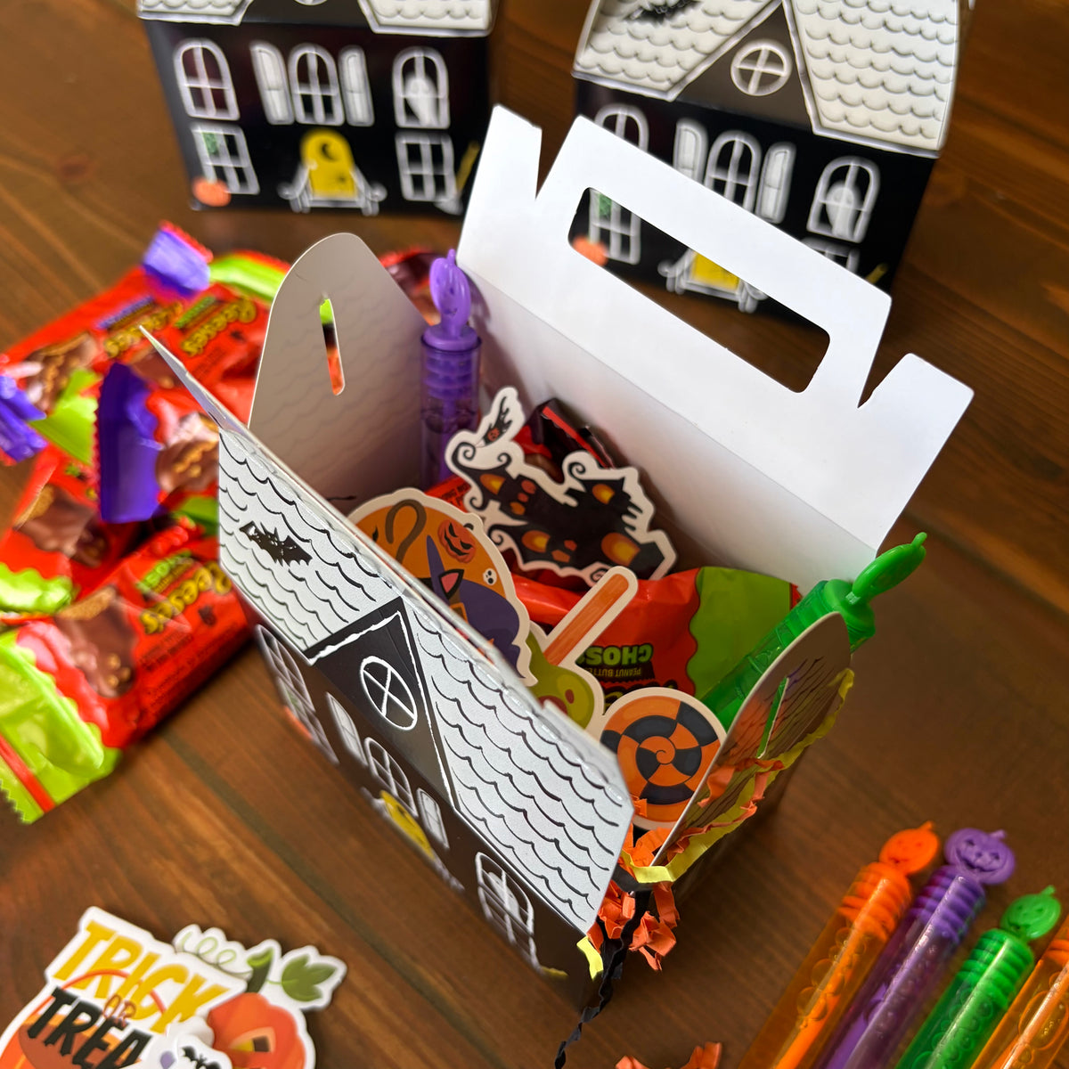 Undecorated white haunted house paper treat box with a carry handle surrounded by crayons and sticker sheets on a wood table