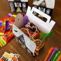 Undecorated white haunted house paper treat box with a carry handle surrounded by crayons and sticker sheets on a wood table
