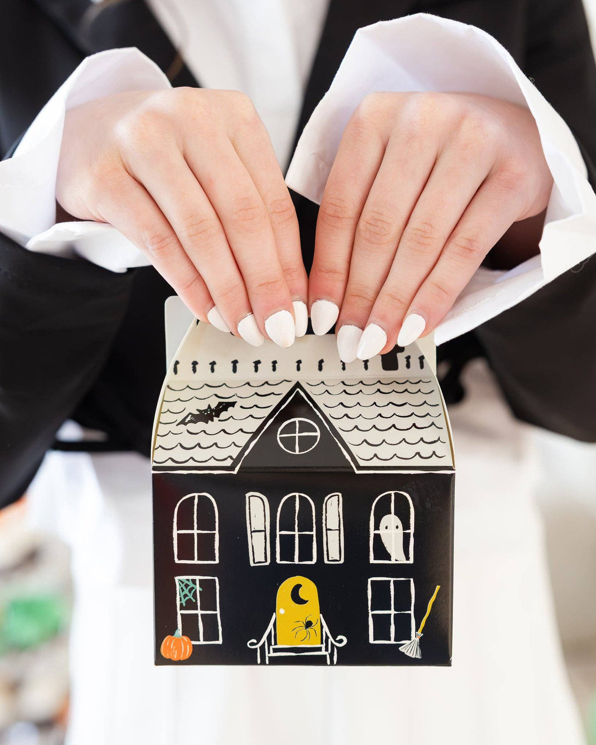Close-up of two hands with white-painted nails lifting a haunted house box by its cut-out handle, showing the cream shingled roof, round attic window, black bat, ghost in a window, yellow glowing door with a crescent moon and spider, orange pumpkin, and broom. Detail shot.