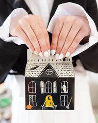 Close-up of two hands with white-painted nails lifting a haunted house box by its cut-out handle, showing the cream shingled roof, round attic window, black bat, ghost in a window, yellow glowing door with a crescent moon and spider, orange pumpkin, and broom. Detail shot.