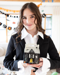 Smiling teen girl in a black blazer and white collared shirt holds one assembled black haunted house treat box at chest height; soft-focus Halloween banners behind her including an orange scalloped swag and a black letter banner. Bright lifestyle portrait.