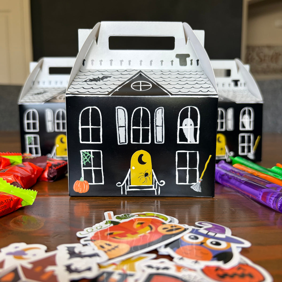Decorated haunted house paper treat boxes with black window stickers, crayons, and pumpkin stickers spread on a wood table