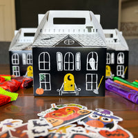 Decorated haunted house paper treat boxes with black window stickers, crayons, and pumpkin stickers spread on a wood table
