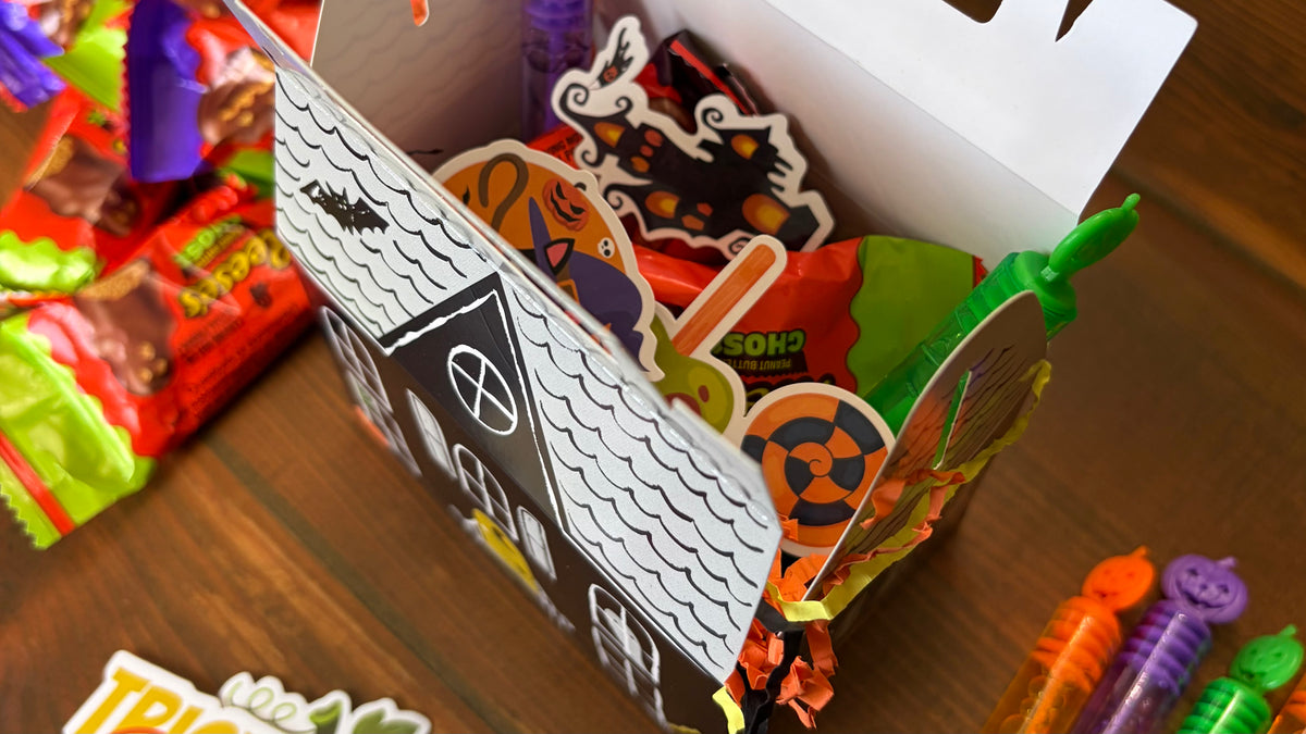 Kids assembling white haunted house paper treat boxes on a wood table with scattered crayons, sticker sheets, and party favors