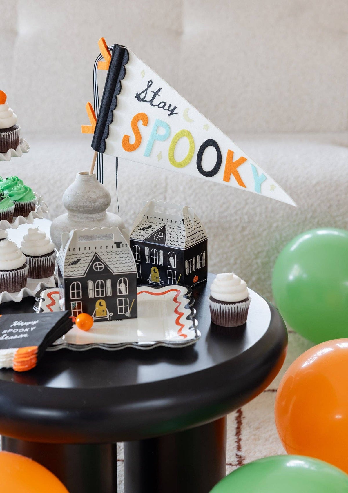 Styled tabletop vignette: a "Stay Spooky" pennant flag rises from a stone vase behind two assembled haunted house boxes; a white scalloped plate with an orange squiggle edge, a stack of candy-corn-edged napkins, chocolate cupcakes, and green and orange balloons fill the frame. Angled close shot.