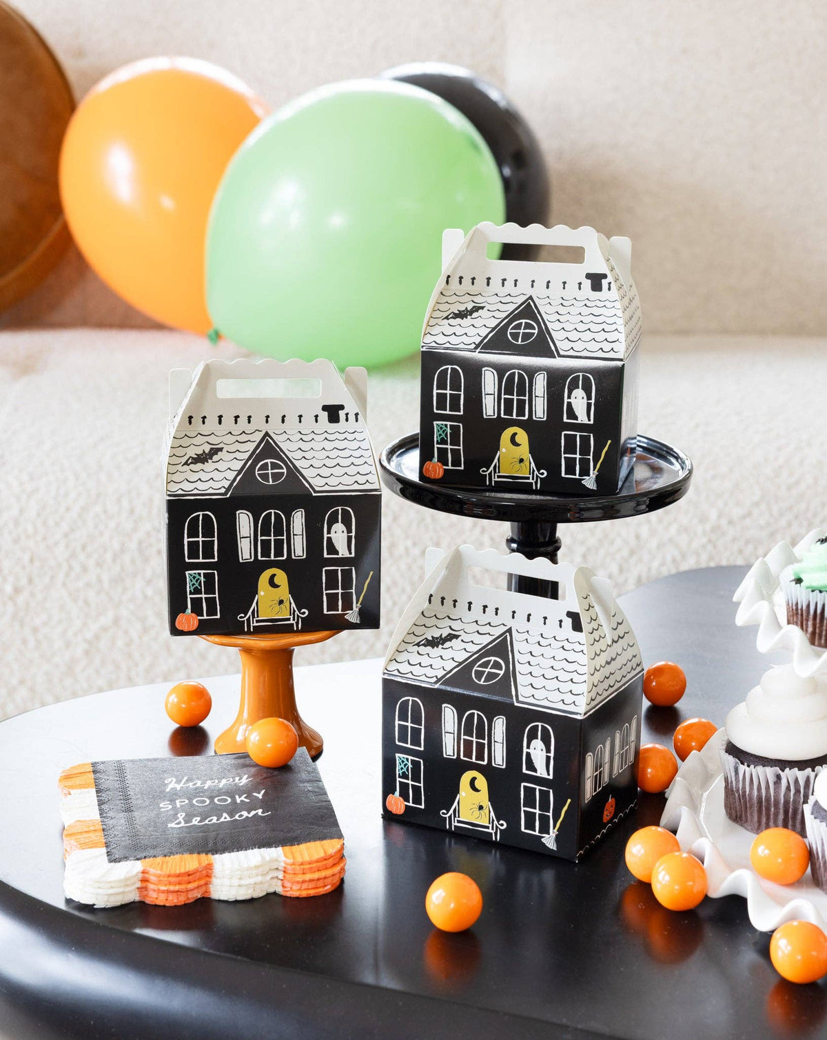 Three assembled haunted house boxes styled at varying heights — one on an orange pedestal, one on a black cake stand, one on the table — alongside candy-corn-edged napkins, orange gumballs, cupcakes, and balloons. Product-in-scene shot.