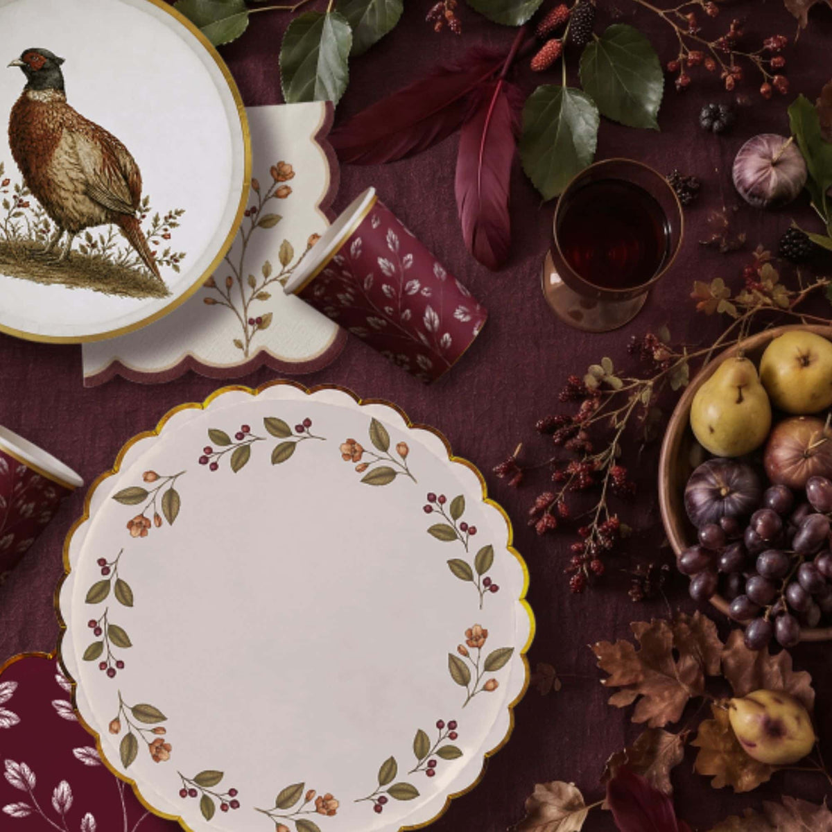 Fall table on burgundy linen with a pheasant plate, berry wreath plate, burgundy leaf cups, and a berry sprig napkin