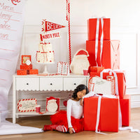 Full shot of a holiday table decor display with a cake, large red gifts, and a girl resting near the setup, featuring the Santa placemats.