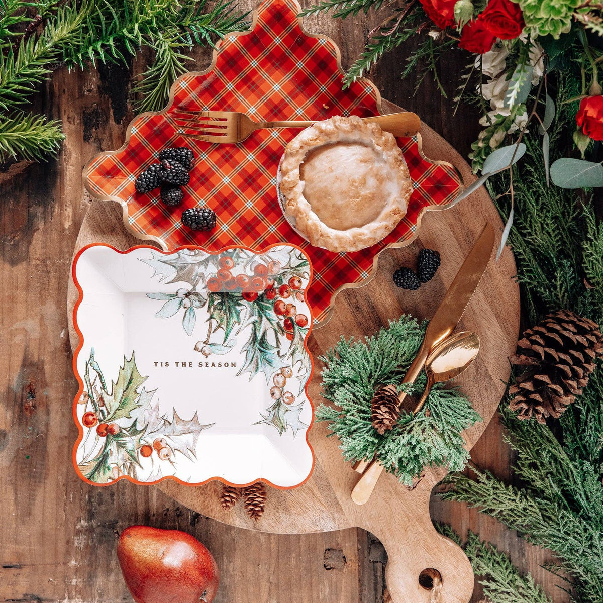 Overhead view of holly and plaid plates styled with berries, gold cutlery, and greenery