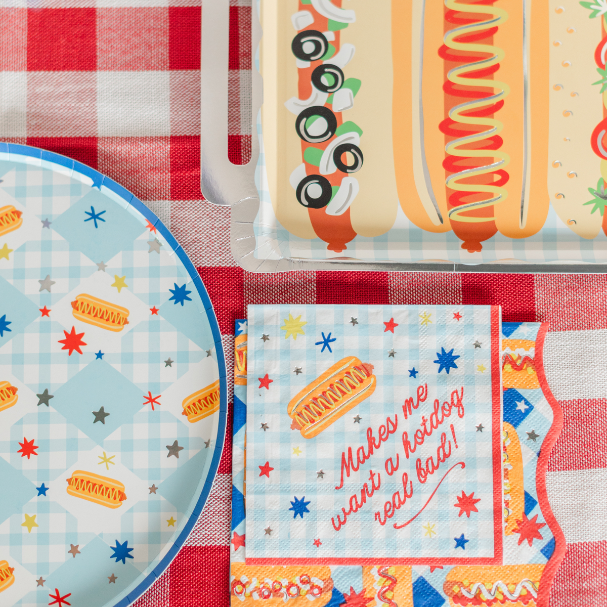 Square hot dog cocktail napkin printed "Makes me want a hotdog real bad!" on red gingham, beside a blue gingham hot dog plate and a foil-edged plate with realistic hot dogs.