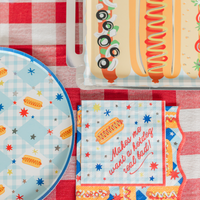Square hot dog cocktail napkin printed "Makes me want a hotdog real bad!" on red gingham, beside a blue gingham hot dog plate and a foil-edged plate with realistic hot dogs.