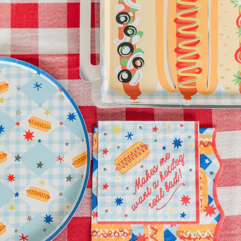 Square hot dog cocktail napkin printed "Makes me want a hotdog real bad!" on red gingham, beside a blue gingham hot dog plate and a foil-edged plate with realistic hot dogs.