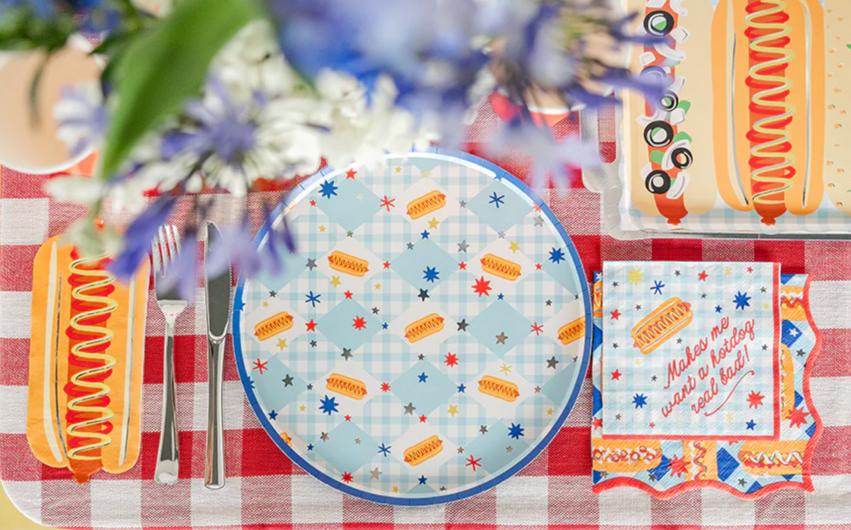 Hot dog party place setting with a blue gingham hot dog plate, silver cutlery, a die-cut hot dog napkin, and the square "Makes me want a hotdog real bad!" cocktail napkin on a red gingham cloth.