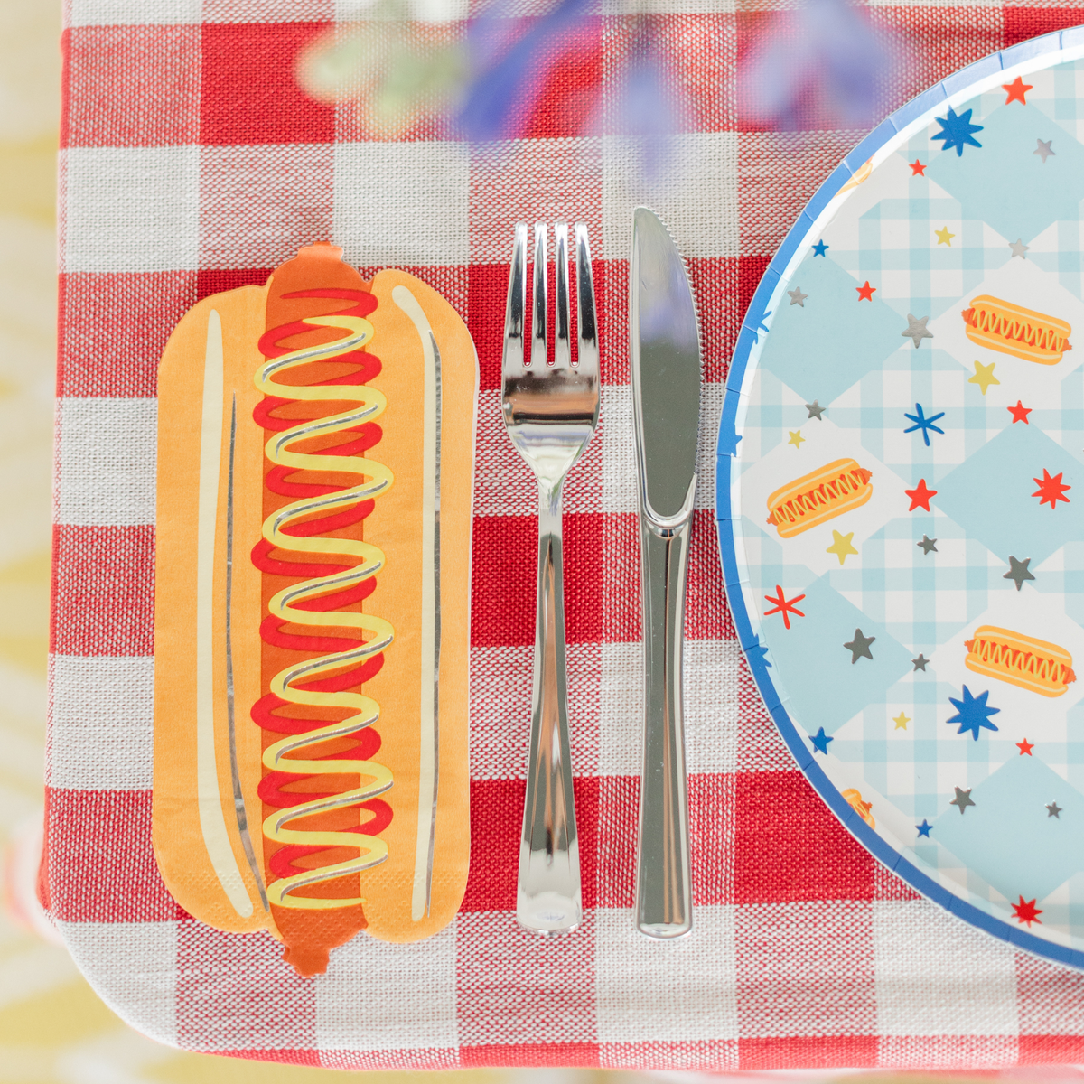 Hot dog shaped paper guest napkin on a red gingham tablecloth beside a silver fork and knife and a blue gingham plate printed with hot dogs and stars.