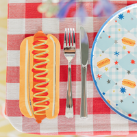 Hot dog shaped paper guest napkin on a red gingham tablecloth beside a silver fork and knife and a blue gingham plate printed with hot dogs and stars.