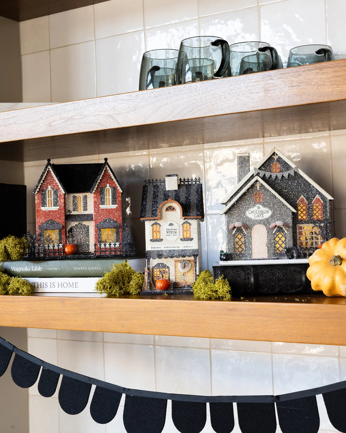 Three glitter Halloween village houses styled on a kitchen shelf with books, moss and a white pumpkin
