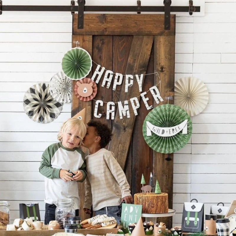A styled kids' camp birthday party scene with teepees, rustic wood decor, paper fans, and a cozy picnic setup under “Happy Camper” signage.